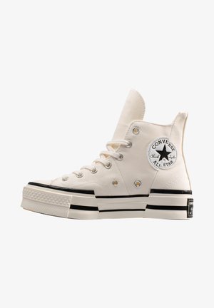Zapatillas de caña alta Chuck Taylor All Star en lona crema con suela de goma negra. Presenta ojetes plateados y un parche de logo negro en forma de estrella.