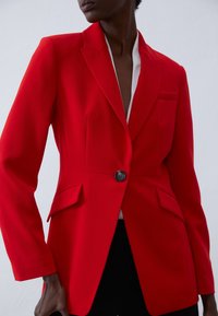 Blazer rouge sur mesure avec une fermeture à un seul bouton, des revers crantés, deux poches avant, et une texture de tissu lisse. Accentué par une doublure intérieure blanche.