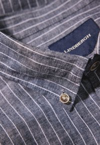 Blauw gestreept overhemd met een geweven textuur, knoopsluiting en een label met "Lindbergh" in het wit op een donkere achtergrond.
