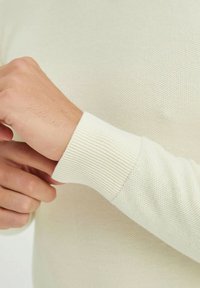 Pull en crème clair avec des poignets côtelés et un corps en tricot texturé. Mettez l'accent sur une main ajustant la manche pour le détail.