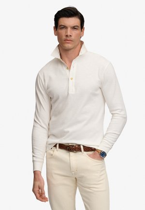 Mand iført cremfarvet langærmet polotrøje med løftet krave, cremefarvede bukser, brunt flettet bælte og armbåndsur med blå urskive, hånd i lommen.