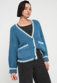 Μπλε αφράτο ζακέτα με V λαιμόκοψη, αντίθετη φωτεινή γαλάζια λεπτομέρεια, δύο εμπρόσθιες τσέπες και καφέ κουμπιά· φορεμένη πάνω από μαύρο τοπ.