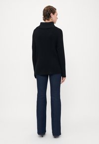 Person som bär en svart rippad polotröja och mörkblå utsvängda jeans, står mot en enkel vit bakgrund, vänd bort.