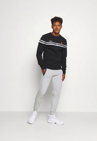 Juodas džemperis su baltais dryžiais ir logotipu, derinamas su pilkomis sportinėmis kelnėmis. Modelis vilki baltus sportinius batelius, stovėdamas priešais paprastą foną.