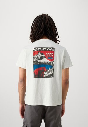Henkilö, jolla on rastat ja valkoinen t-paita, jossa on takana punainen, sininen ja musta grafiikka karhusta, järvestä ja vuoresta, merkintänä "NAPAPIJRI 1987".