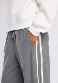 Lichtgrijze sweatshirt met ribgebreide manchetten en een witte trekkoord. Grijze broek met zijzakken en witte strepen langs de zijkanten.