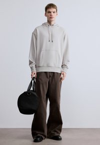 Helehall kapuutsiga dressipluus ees taskuga, kombineeritud pruunide laiade pükstega. Modell hoiab ümarat musta käepidemega kotti.