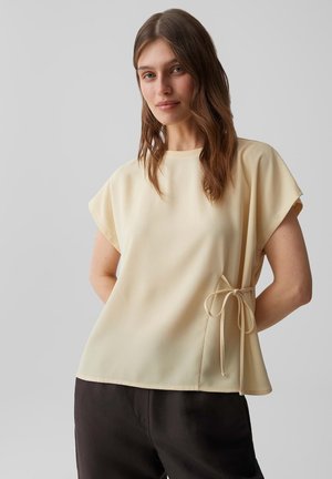 Donna con lunghi capelli castani che indossa una maglietta crema a maniche corte con fiocco laterale e pantaloni neri, in piedi davanti a uno sfondo grigio uniforme.
