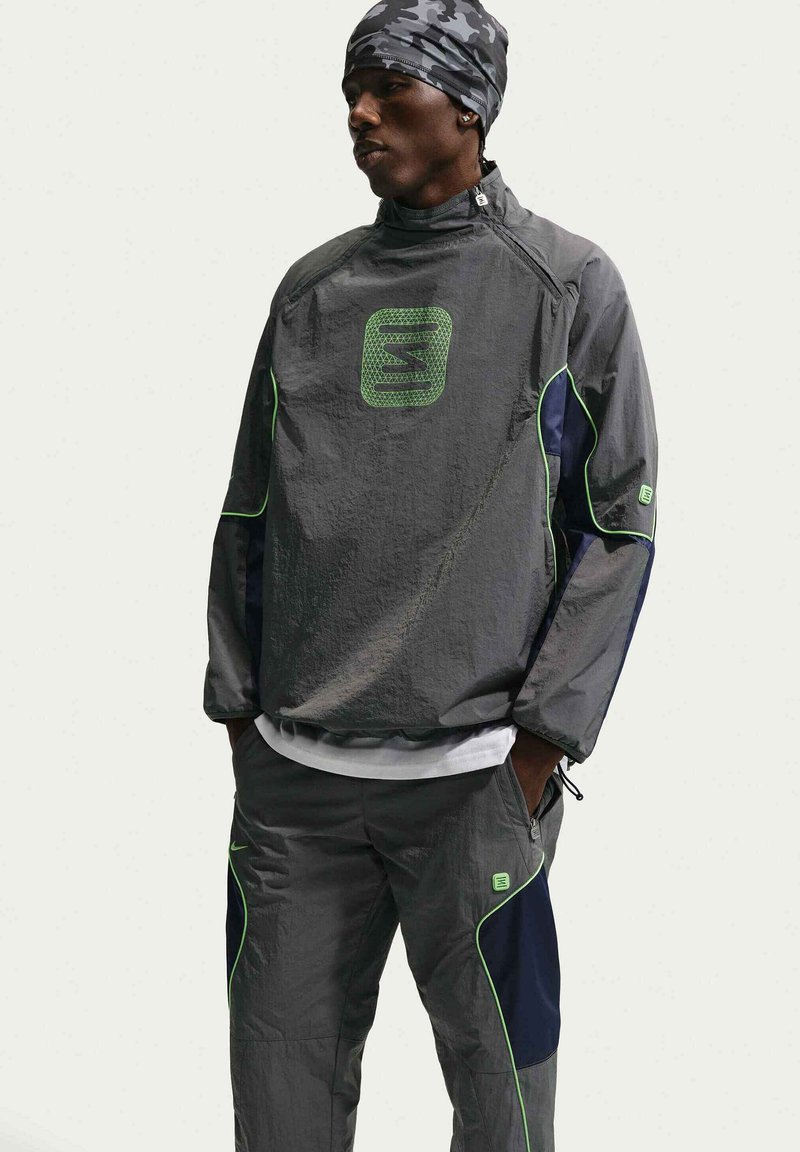 Man in een grijs trainingspak met groene accenten en een camouflage hoofdbedekking, staat met de handen in de zakken en kijkt naar links.