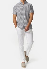 Man in een grijs-witte verticaal gestreepte overhemd met korte mouwen, witte broek en beige instappers die naar voren loopt.