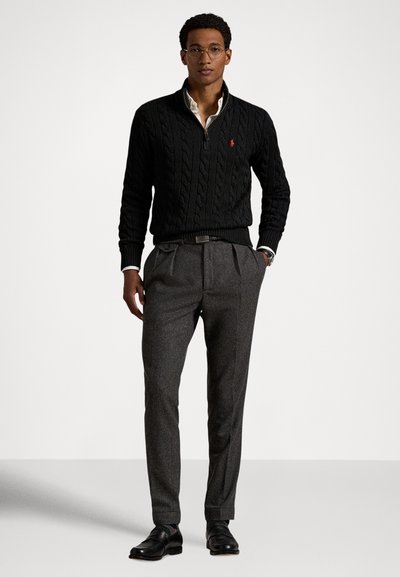 Μαύρο πουλόβερ με πλέξη καλώδιο και φερμουάρ στο μισό, γκρι πλισέ παντελόνια, μαύρα παπούτσια και γυαλιά. Λευκό γιακά πουκάμισου φαίνεται κάτω από το πουλόβερ.