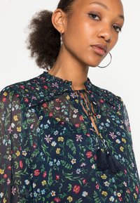Blusa floreale in tessuto blu navy trasparente, con colletto volant, apertura a chiave con lacci e dettagli con nappine. Piccole fiori multicolori su tutta la superficie.
