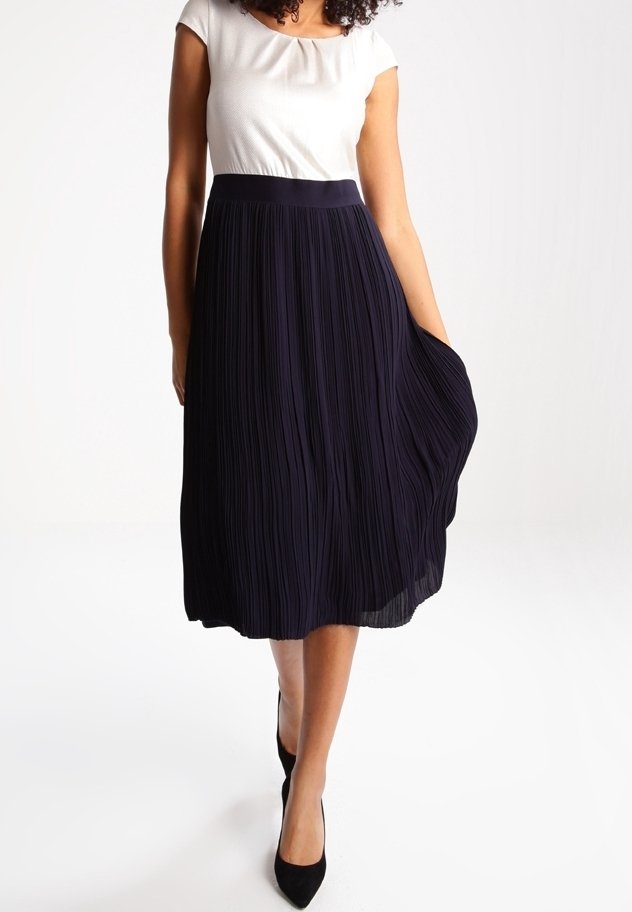 Femme portant un haut blanc à manches courtes rentré dans une jupe midi plissée bleu marine taille haute et des chaussures noires pointues.