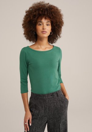Haut vert à manches longues avec un décolleté rond, présentant une texture lisse, associé à un pantalon gris à taille haute et à motifs.