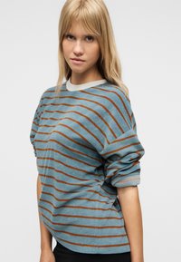 Νεαρή γυναίκα με μακριά ξανθά μαλλιά που φοράει μπλε πουλόβερ με καφέ οριζόντιες ρίγες, στέκεται μπροστά σε ένα απλό φωτεινό φόντο.