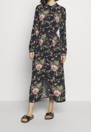 Femme portant une robe longue à manches longues bleu marine à fleurs roses et vertes, taille nouée, fente à l'avant, et sandales plates noires avec boucles.