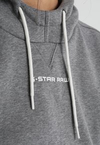 Sudadera gris con cuello alto, que presenta cordones blancos y un logo impreso "G-STAR RAW". Textura suave, diseño casual.