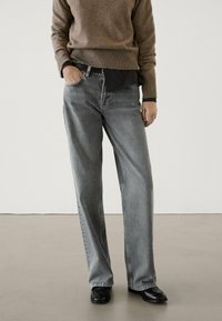 Jeans larghi grigi con una finitura sbiadita, che presentano un classico design a cinque tasche, passanti per cintura e dettagli di cucitura sottili, abbinati a un maglione marrone.