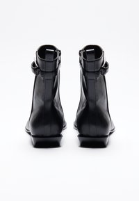 Bottines en cuir noir présentant un design épuré, des zippers latéraux et un petit talon carré. Les accents incluent des sangles ajustables au niveau du haut.