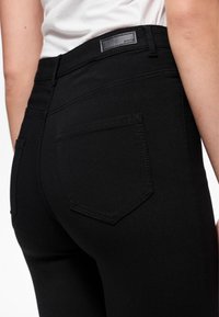 Jean en denim noir extensible avec une taille haute, doté d'un patch en cuir sur la ceinture et de deux poches arrière, avec des détails de couture subtils.
