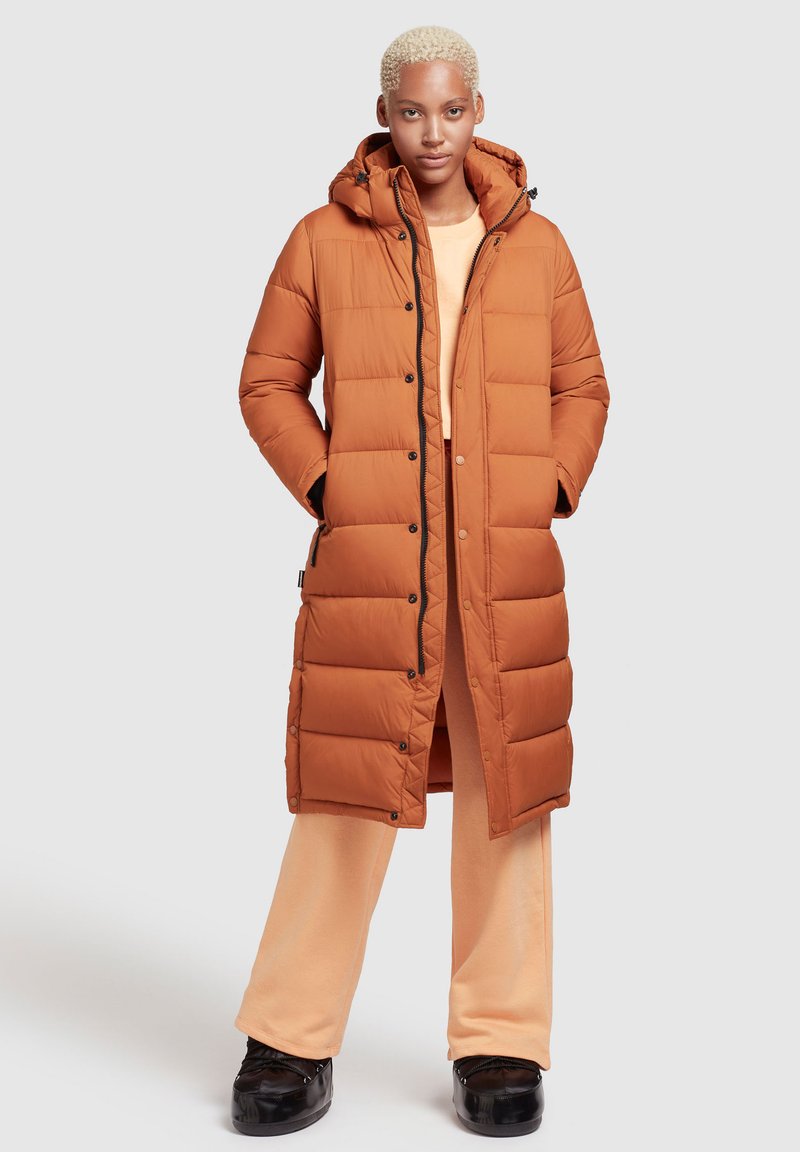 Langer orangefarbener Daunenmantel mit Kapuze, ausgestattet mit einem gesteppten Design, schwarzen Knöpfen und seitlichen Taschen, getragen über einem hellorangefarbenen Outfit.