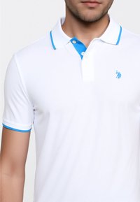 Kortærmet hvid polo med blå krave og manchetter, prydet med et lille logo på brystet. Modellen viser skjorten mod en lys baggrund.