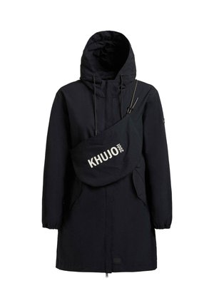 Schwarzer langer Kapuzenmantel mit elastischen Bündchen und großer schwarzer Umhängetasche mit der Aufschrift "KHUJO 2001" quer über der Brust.