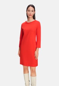 Rotes, langärmliges Kleid mit Rundhalsausschnitt und knielangem Design, aus glattem Stoff gefertigt. Kombiniert mit hellen, kniehohen Stiefeln.