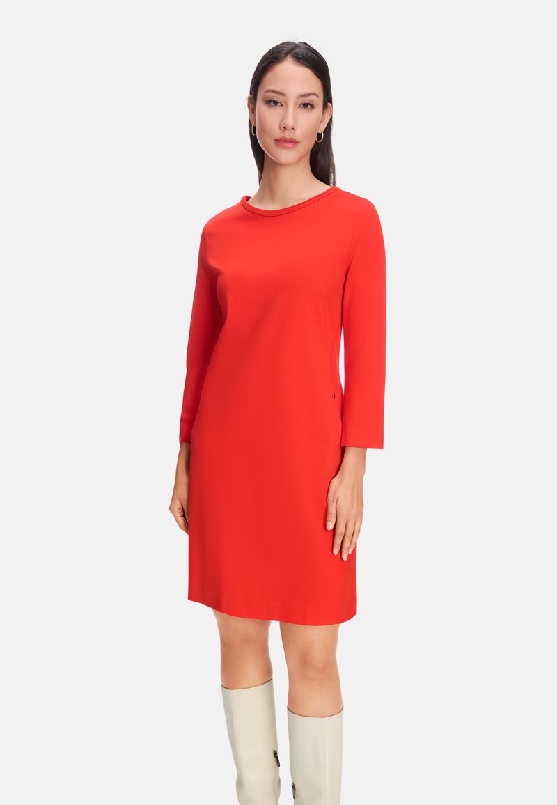 Rotes, langärmliges Kleid mit Rundhalsausschnitt und knielangem Design, aus glattem Stoff gefertigt. Kombiniert mit hellen, kniehohen Stiefeln.
