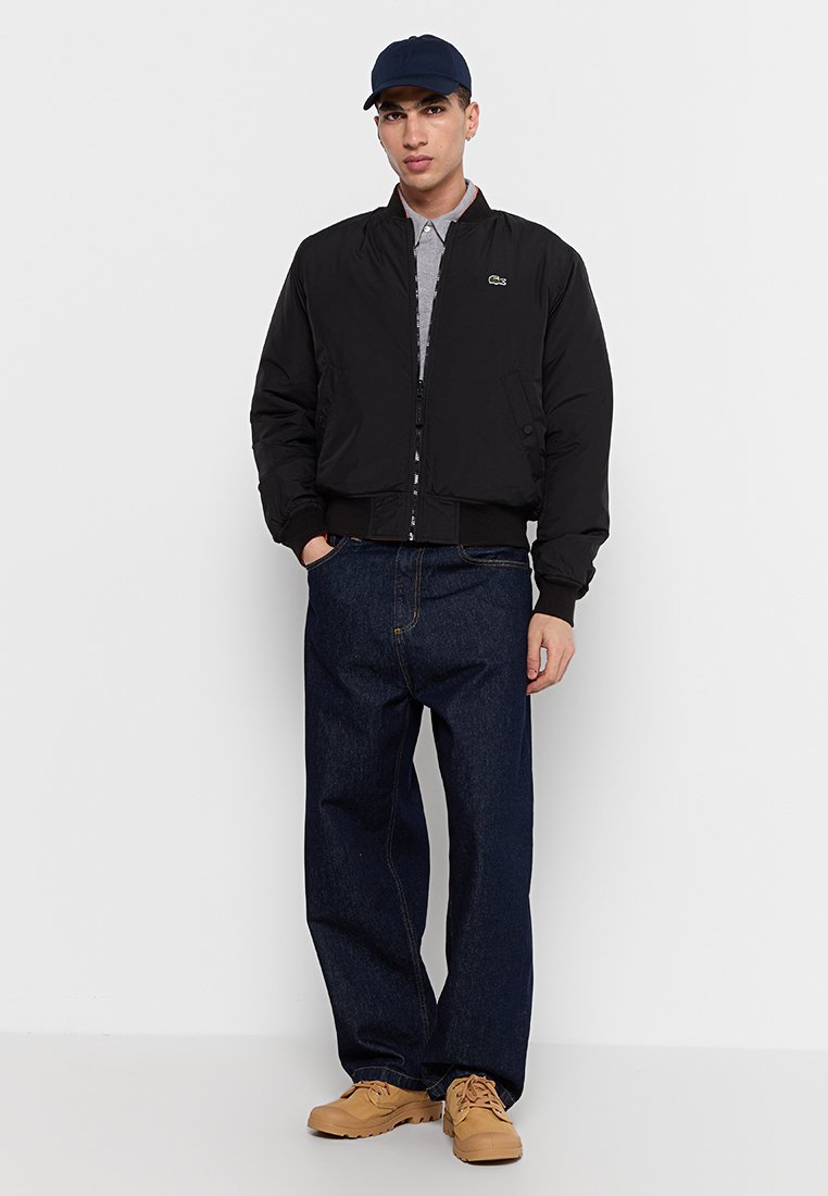 Veste bomber noire avec des poignets côtelés, poches latérales à boutons, associée à une chemise grise à col et un jean baggy bleu foncé. Des chaussures caramel complètent le look.