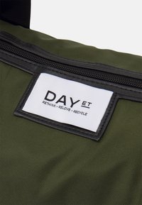 Grön tygväska med strukturerad yta, utrustad med svart dragkedja och en vit etikett med texten "DAY ET" och återvinningslogotyper.