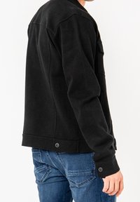 Sort, tekstureret jakke med lige snit, der har høj krave og knapdetaljer, båret over blå jeans.