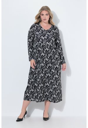 Robe noire à manches longues avec un motif de feuilles blanches, cintrée à la taille, jupe fluide, col rond, portée avec des talons noirs.