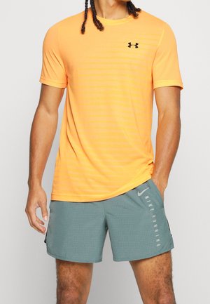 Męski model noszący jaskrawopomarańczowy krótki rękaw Under Armour i jasnoszare spodenki Nike Running, stojący z jedną ręką w kieszeni.