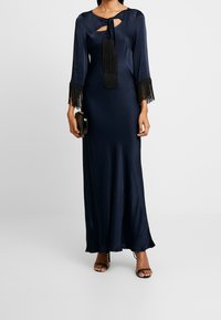 Vestido largo de satén marino con mangas largas, escote en forma de llave y detalles de flecos negros en los puños. La modelo sostiene un bolso negro y lleva sandalias de tiras.