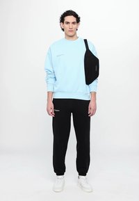Lyseblå sweatshirt med rund hals, sorte sweatpants og hvide sneakers. Sort crossover-taske på venstre skulder. Tekstdetalje på sweatshirten.