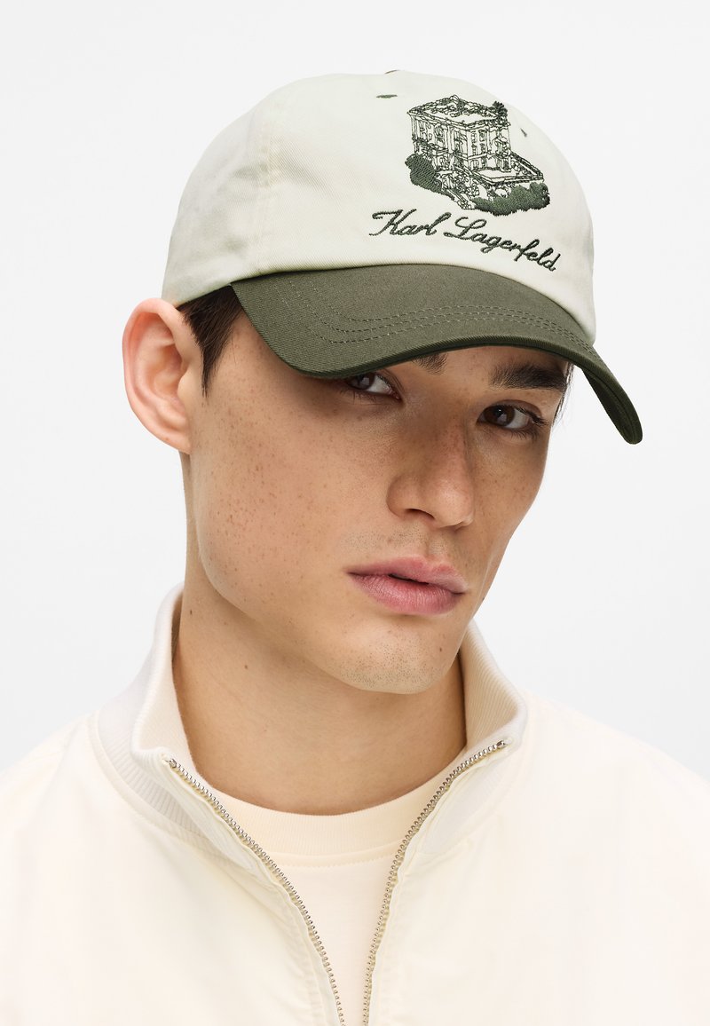 Gorra beige con visera verde, presenta un edificio bordado y el texto "Karl Lagerfeld". Fabricada con tela de textura suave.