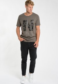 Olive green t-shirt with a black ornate letter "N" graphic, paired with black jeans and white sneakers. Set against a plain background.