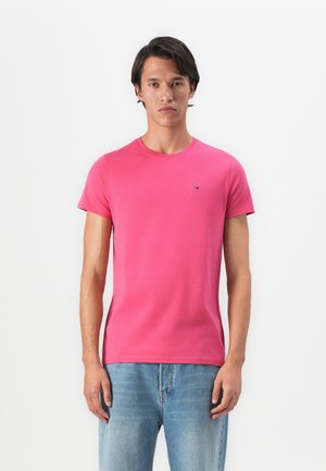 Un jeune homme debout, portant un t-shirt à manches courtes rose vif et un jean bleu clair, tourné vers l'avant devant un fond blanc uni.