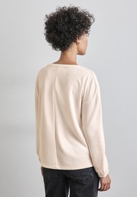 Beige Sweatshirt mit langen Ärmeln, rundem Halsausschnitt und lässiger Passform. Verfügt über dezente Nähte und elastische Bündchen, kombiniert mit dunklen Jeans.