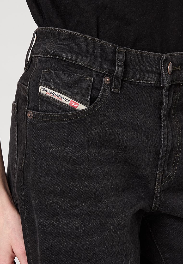 Jeans in denim nero indossati da una persona, che mostrano una tasca con l'etichetta bianca e rossa della Diesel Industry ben visibile.