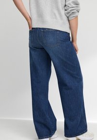 Weite blaue Jeans aus Denim haben eine hohe Taille, strapazierfähigen Stoff und zwei Gesäßtaschen. Das Modell trägt einen grauen Pullover und helle Sneakers.