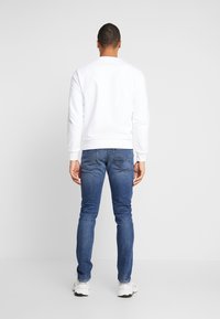 Biała bluza z okrągłym dekoltem noszona z dopasowanymi niebieskimi dżinsami. Bluza ma gładką fakturę, a dżinsy cechują się klasycznym wykończeniem z denimu.