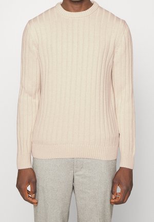 Mężczyzna ubrany w jasnobeżowy prążkowany sweter z dzianiny i jasnoszare spodnie, stojący na tle jednolitej białej ściany.