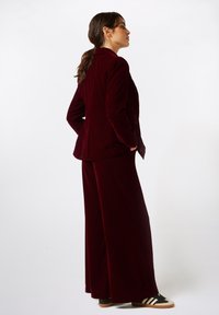 Traje de terciopelo burdeos con pantalones de pierna ancha y blazer ajustado, combinado con zapatillas a rayas en blanco y negro. Textura suave, ajuste a medida.