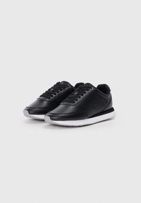 Un paio di sneakers nere basse con suole bianche, lacci neri e logo "cK" sui lati, mostrato su uno sfondo leggero e semplice.