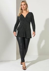 Czarna, błyszcząca bluzka z głębokim dekoltem w serek i długimi rękawami, zestawiona z czarnymi, dopasowanymi spodniami ze sztucznej skóry i cienkimi, paskowymi szpilkami, trzymająca srebrną kopertówkę.