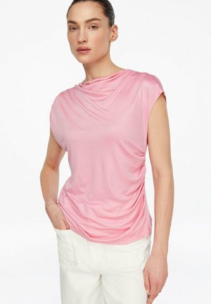 Kobieta z ciemnymi włosami, ubrana w różową bluzkę z krótkim rękawem i białe spodnie, stojąca z jedną ręką w kieszeni na białym tle.