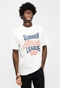 Λευκό βαμβακερό μπλουζάκι με έντονο κόκκινο και ναυτικό μπλε κείμενο "SUMMER Athletic LEAGUE" με το λογότυπο της Adidas, σε συνδυασμό με μαύρο τζιν.