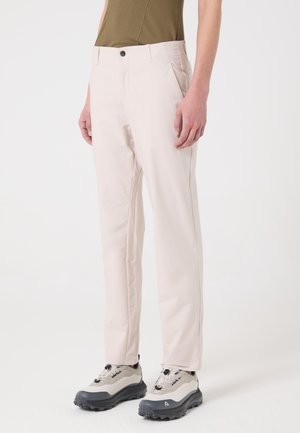 Person steht und trägt hellbeige gerade geschnittene Hosen, ein braunes strukturiertes Oberteil und beige Wanderschuhe mit schwarzen Sohlen vor einem schlichten weißen Hintergrund.